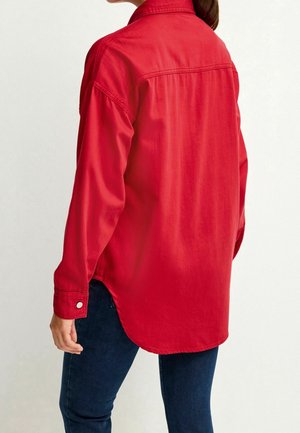 Nainen, jolla on yllään löysä punainen pitkähihaisuus napillinen paita kaarevalla helmalla ja tummansiniset farkut, kääntyy poispäin kamerasta.