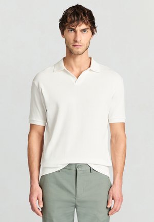 Homme aux cheveux bruns ondulés portant un polo blanc à manches courtes côtelé et un pantalon vert clair, debout devant un fond uni.