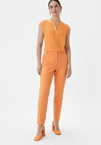 Orange sleeveless top with a V-neck and matching slim-fit trousers. The outfit features a smooth texture and is paired with orange block-heeled shoes.
