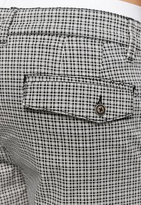 Schwarz-weiße Karostoff mit einer hinteren Tasche mit Knopfverschluss. Die Textur ist glatt und gewebt, wodurch ein Rautenmuster entsteht.
