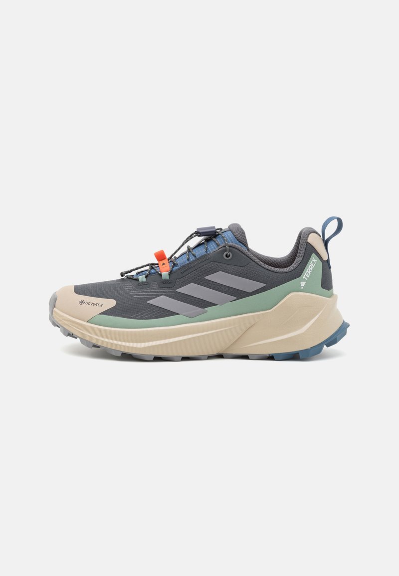 Shoe de corrida em trilha em cinza escuro, com uma entressola bege e detalhe verde. Apresenta um cabedal texturizado, um sistema de atacadores ajustável e uma sola robusta.