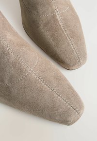 Beige mokkasiininsyöiset nilkkasaappaat, joissa on terävä kärki, näkyvät ompelut yksityiskohdat ja sileä pinta. Suunnittelu on tyylikkään moderni siluetti.