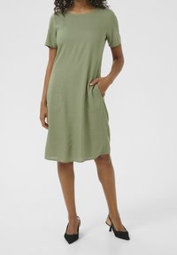 Vrouw in een olijfgroen jurk tot op de knie met korte mouwen en zakken, gecombineerd met zwarte slingback hakken met puntige neuzen, staande op een witte achtergrond.