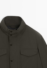 Giacca verde scuro con colletto alto, presenta chiusure frontali con bottoni e due tasche sul petto con dettagli a pattina. Tessuto dalla consistenza liscia.