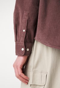 Gros plan sur la main gauche d’une personne à côté d’une poche de pantalon beige, portant une chemise marron en velours côtelé à manches longues avec des poignets boutonnés.