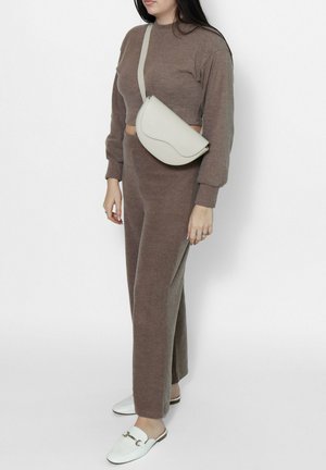 Vrouw die een bruin gebreid tweedelig outfit draagt, met een witte crossbody tas en witte instap loafers, staand tegen een effen witte achtergrond.