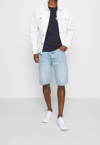 Chaqueta de mezclilla blanca sobre una camiseta de color navy, pantalones cortos de mezclilla azul claros y zapatillas deportivas blancas. Presenta dos bolsillos frontales y un estilo casual.