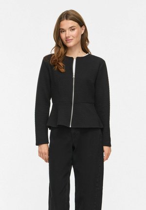 Femme aux longs cheveux bruns portant une veste noire texturée zippée avec un ourlet péplum et un pantalon noir, debout sur un fond blanc uni.