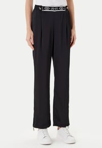 Pantaloni neri in jersey con un design plissettato, vita elasticizzata con il marchio "LIU JO" e dettagli con zip all'orlo. Indossati con sneakers bianche.