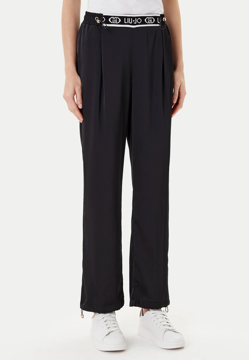 Pantaloni neri in jersey con un design plissettato, vita elasticizzata con il marchio "LIU JO" e dettagli con zip all'orlo. Indossati con sneakers bianche.