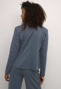 Modrý blazer s vzorem, dlouhými rukávy a jemně strukturovaným střihem, s geometrickým designem a texturovanou tkaninou, zobrazený ze zadní strany.