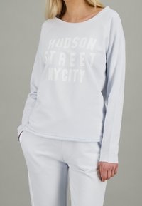 Sudadera de color azul claro con "HUDSON STREET NYC" en letras blancas, mangas largas, puños acanalados y un corte relajado. Textura de tela suave.