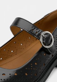 Μαύρο παπούτσι από δέρμα βινυλίου με διάτρητο σχέδιο, στρογγυλεμένη μύτη και ρυθμιζόμενο λουράκι με μεταλλική αγκράφα. Λεπτή εσωτερική επένδυση.