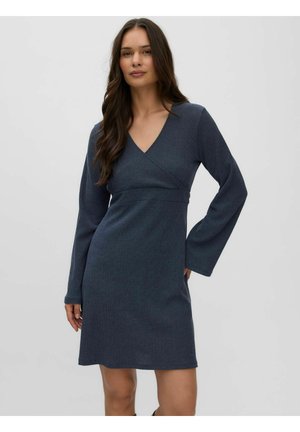 Femme aux longs cheveux bruns portant une robe bleu foncé à manches longues et col en V, debout devant un fond clair uni.
