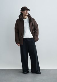 Veste marron rembourrée avec capuche, sweat-shirt blanc, pantalon de survêtement large noir et chaussures noires. Accessoires comprenant une casquette marron assortie.
