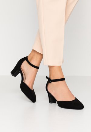 Zwarte suède hoge hakken met een enkelband, ronde neus en geometrische blokhak, gedragen met lichte broek.