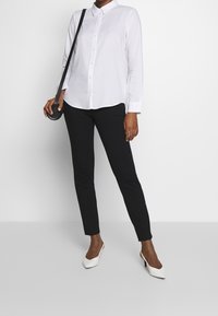 Weißes langärmeliges Hemd mit Knöpfen, schwarze eng anliegende Hose und weiße Pumps. Die Accessoires umfassen eine kleine schwarze Umhängetasche.