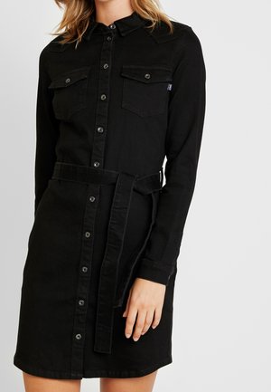 Kobieta ubrana w czarną sukienkę jeansową na guziki z długimi rękawami, kieszeniami z klapkami na piersi oraz wiązanym materiałowym paskiem w talii.