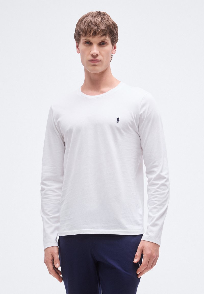 Hvit langermet T-skjorte i bomull med rund hals og en liten marineblå logo på venstre bryst. Modellen har på seg marineblå bukser.