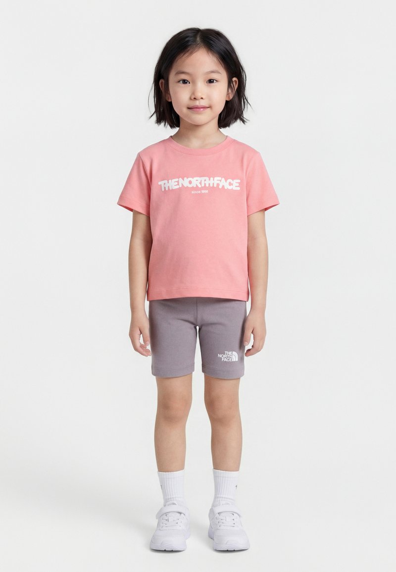 Νεαρή κοπέλα που στέκεται, φοράει ροζ μπλούζα The North Face, γκρι σορτς, λευκές κάλτσες και λευκά αθλητικά παπούτσια μπροστά σε απλό φόντο.