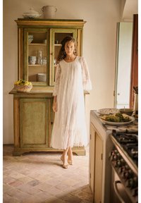 Žena v dlhých bielych čipkovaných šatách stojí v rustikálnej kuchyni vedľa zelenej drevenej skrinky a koša s citrónmi na dlažobnej podlahe.