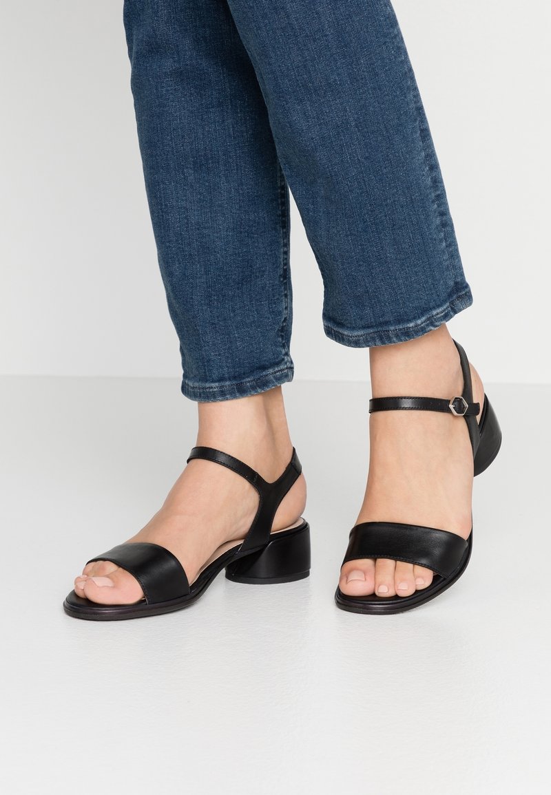 Mustat nahkaiset korolliset sandaalit, joissa on leveät varvasremmit, nilkkaremmi ja paksu korko. Suunnittelussa on minimalismi ja sileä pinta.