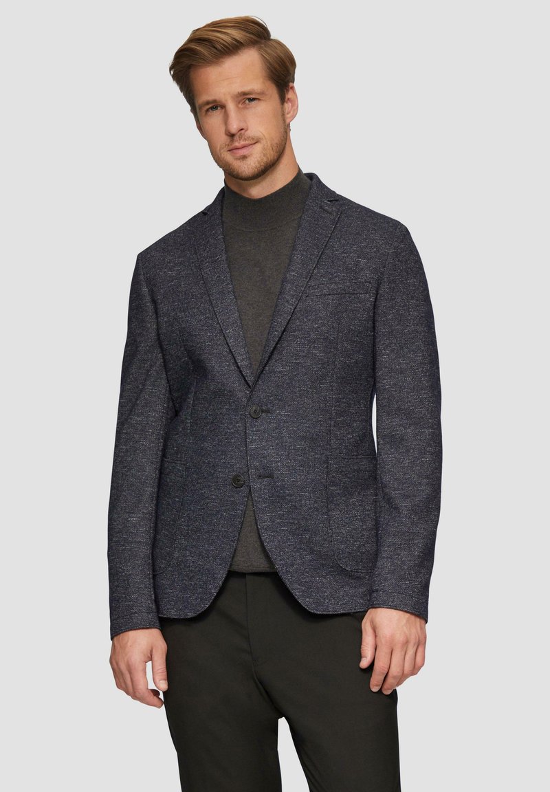 Maßgeschneiderter marineblauer Blazer aus strukturiertem Stoff, mit einer Zwei-Button-Front, Reverskragen und Klappentaschen. Kombiniert mit einem dunklen Rollkragenpullover darunter.