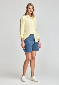 Ljusgul linne skjorta med knappar framtill, blå denim shorts och vita sneakers. Skjortan har uppkavlade ärmar; shortsen är knälånga.