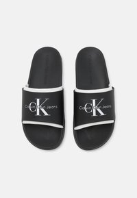 Čierne šľapky s plochým dizajnom, syntetický materiál a biele akcenty s logom "Calvin Klein Jeans" v kontrastnom písme.