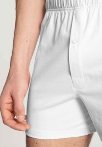 Pantaloni boxer bianchi in cotone con un elastico in vita e chiusura a bottone anteriore, caratterizzati da una texture liscia e un taglio dritto.