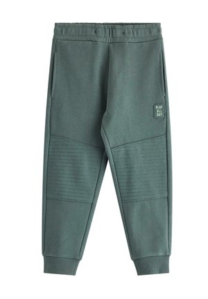 Lyshåret barn iført en brun kasket, stribet langærmet trøje, blå joggingbukser og sneakers, stående i en legesyg positur på en hvid baggrund.