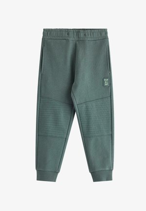 Blondes Kind mit brauner Mütze, gestreiftem Langarmshirt, blauen Jogginghosen und Sneakers, das in einer spielerischen Pose vor weißem Hintergrund steht.