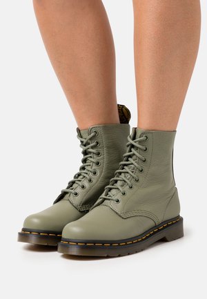 Personne portant des bottines en cuir vert clair à lacets, avec des semelles noires et des coutures jaunes, sur fond blanc.