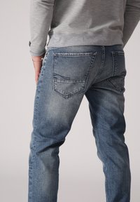 Modré džínové kalhoty se vybledlým povrchem, designem s pěti kapsami a viditelným švem. Zadní pohled ukazuje dvě zadní kapsy se šikmými klapkami.