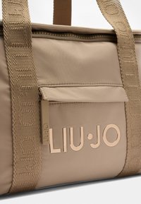 Borsa tote in nylon beige con tasca con zip, parte superiore piatta e dettaglio del logo in oro metallico. Presenta un manico a spalla testurizzato.
