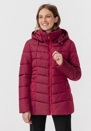 Mujer sonriente que lleva una chaqueta acolchada burdeos con capucha y cremallera, sosteniendo un cordón contra un fondo claro y liso.