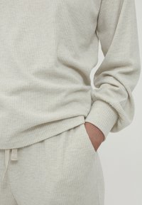 Person trägt ein cremefarbenes, geripptes Langarmoberteil und dazu passende Hosen, mit einer Hand in der Tasche, wobei die Stofftextur und der lässige Schnitt zur Geltung kommen.