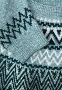 Pull en maille bleu clair et blanc, présentant des motifs en zigzag et en losange en bleu sarcelle foncé. Encolure arrondie, poignets côtelés et texture douce.