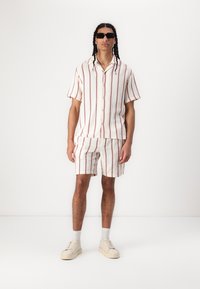 Kortærmet, skjorte med krave og matchende shorts i hvid med røde vertikale striber, lavet af struktureret stof; parret med hvide sneakers.