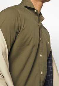 Camicia button-up verde oliva con colletto rialzato, caratterizzata da accenti beige chiaro e una fodera interna a quadri. Tessuto morbido, vestibilità su misura.