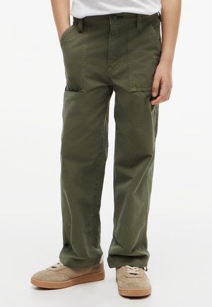 Child wearing olive green cargo pants and beige sneakers, hands in pockets, standing against white background.