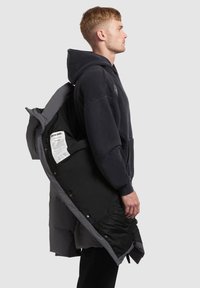 Fekete puffdzseki szürke béléssel, patentos záródással, kapucnival és látható címkével belül. Egy modellel, aki hátizsákot visel.