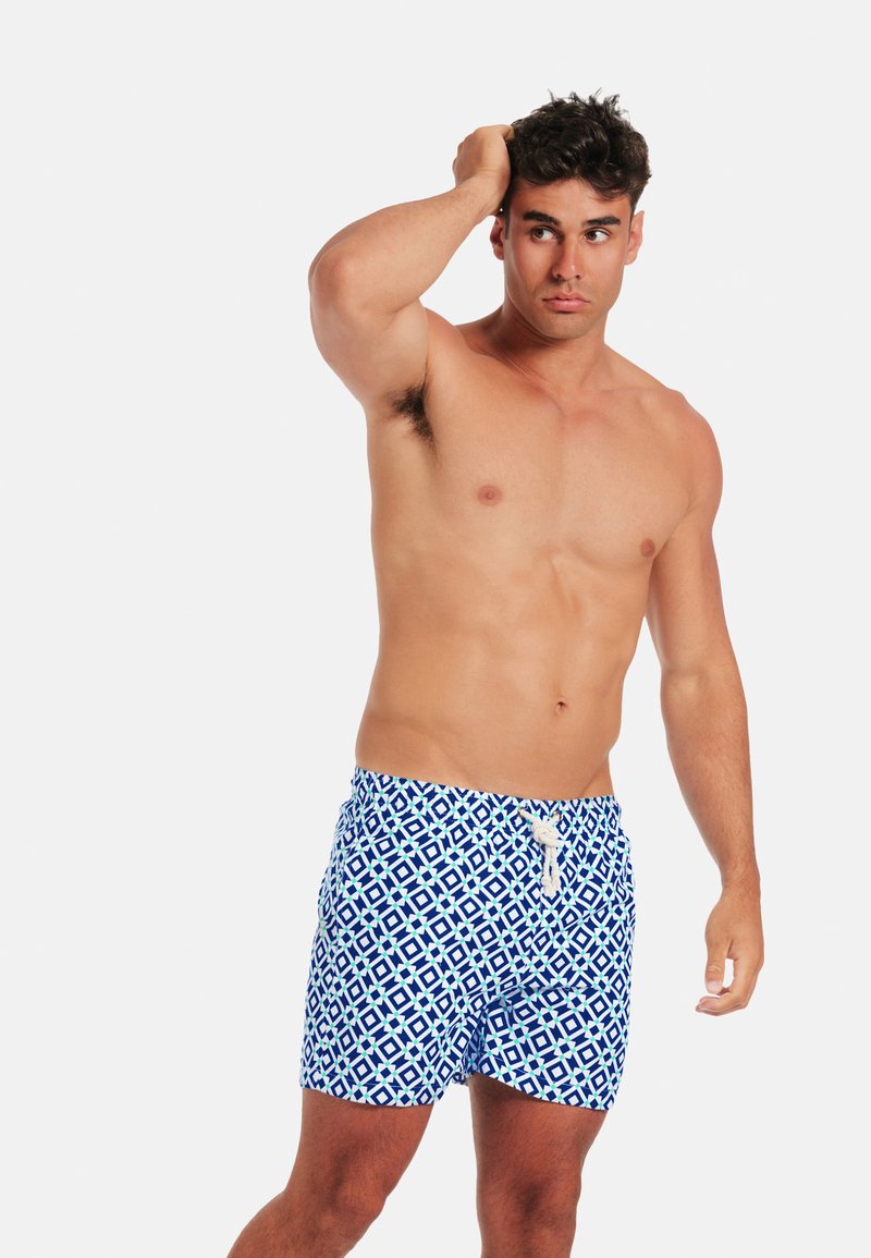 Homme torse nu aux cheveux courts et bouclés portant un short de bain bleu et blanc à motifs, se touchant la tête et regardant sur le côté.