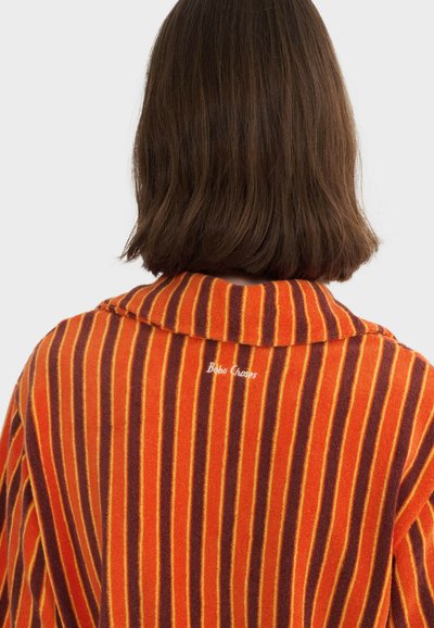 Personne aux cheveux bruns mi-longs vue de dos, portant un vêtement à rayures verticales orange, marron et jaune avec l'inscription « Bobo Choses » brodée au dos.