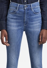Jeans de mezclilla azul con un diseño ajustado, que cuentan con cinco bolsillos, un cierre de botón y un ligero desgaste para un aspecto envejecido. Textura suave.