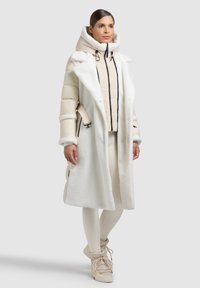 Prošívaná bunda a dlouhý kabát v krémové a bílé barvě. Texturovaná, nadýchaná svrchní vrstva s odnímatelným páskem a kapucí. Kalhoty úzkého střihu a boty.