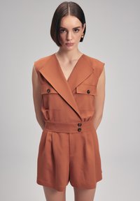 Oranger kurzer Jumpsuit mit tiefem V-Ausschnitt, zwei vorderen Taschen und einem taillierten Gürtel mit Knopfakzenten. Hergestellt aus leichtem Stoff.