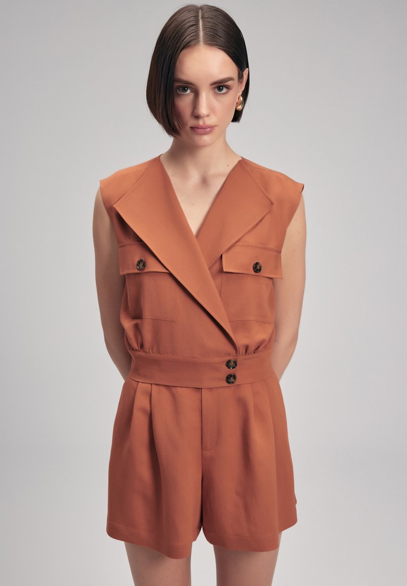 Oranger kurzer Jumpsuit mit tiefem V-Ausschnitt, zwei vorderen Taschen und einem taillierten Gürtel mit Knopfakzenten. Hergestellt aus leichtem Stoff.
