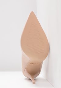 Chaussure à talon haut beige à bout pointu, inclinée vers le haut, montrant une semelle lisse avec une légère texture antidérapante et un petit talon aiguille.