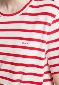 Chemise blanche à rayures rouges horizontales et à manches courtes, arborant le petit logo "GANT" brodé sur la poitrine.